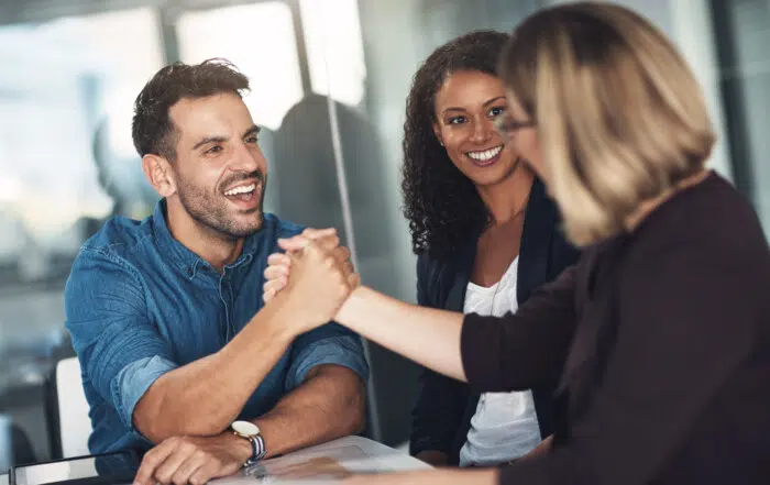 Two people smiling and shaking hands with a third person smiling in the back.